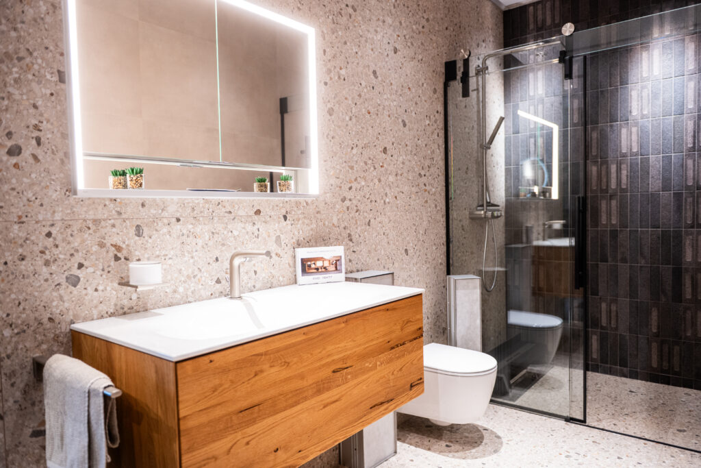 Modernes Badezimmer mit Terrazzo-Fliesen, Holzwaschtisch und Glasdusche bei MABELA Fliesen und Handels GmbH – stilvolle Badgestaltung im Schauraum.